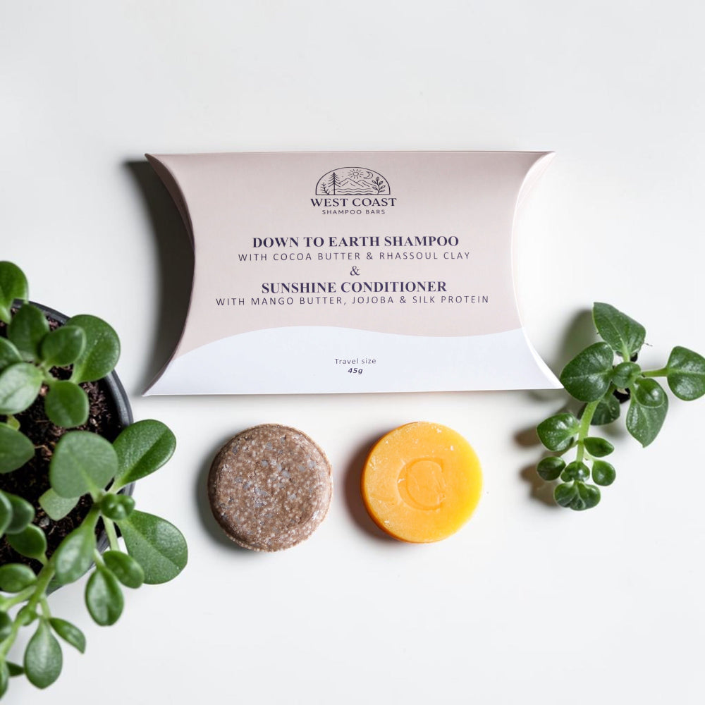 West Coast Cleanse Down to Earth Shampoo and Sunshine Conditioner travel case in blush pink and white, displayed on a white background with two round bars of shampoo (one brown, one yellow) and green plants.