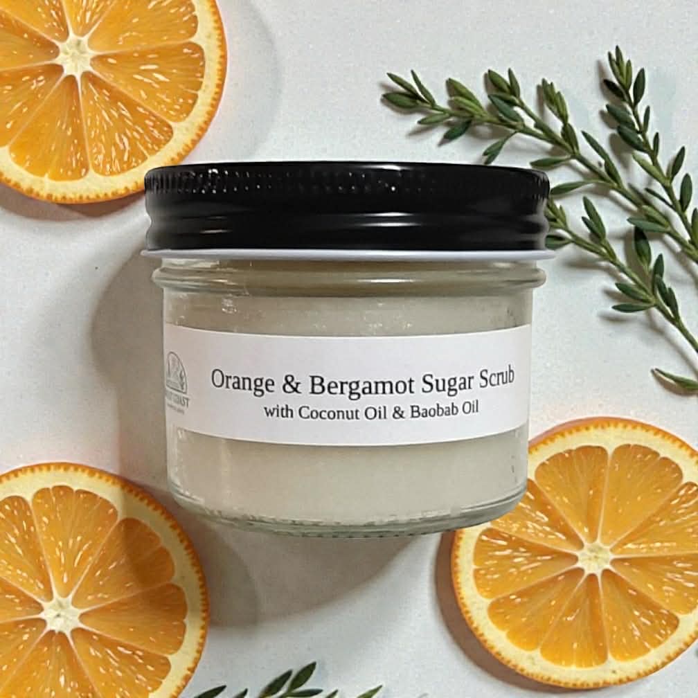 Jar of Orange & Bergamot Sugar Scrub with slices of orange and green leaves on a white background